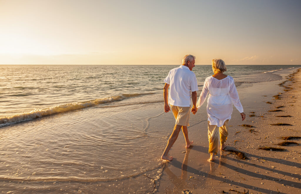 Happy senior man and woman old retired couple walking and holding hands on a beach at sunset, seniorlife