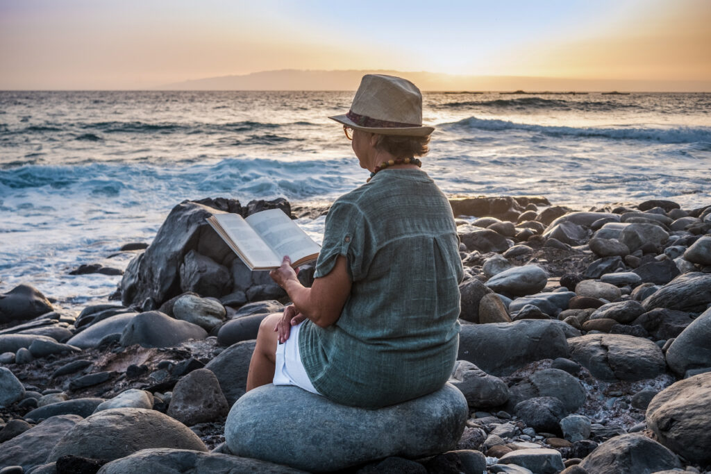 Rear view of alone senior woman with hat sitting on the beach of pebbles reading a book. Waves of the ocean. Sunset light