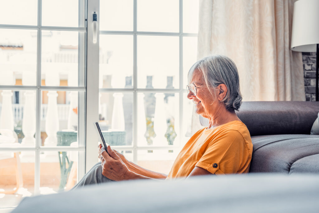 Smiling middle-aged Caucasian woman sit on couch in living room browsing wireless Internet on tablet, happy modern senior female relax on ground at home using pad device, elderly technology concept