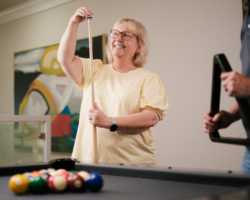 A smiling woman dusts up a pool cue in a well-appointed retirement community.